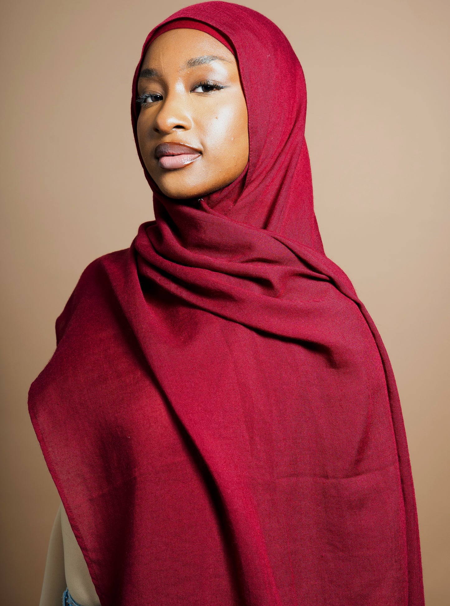 Femme portant un hijab rouge bordeaux en tissu léger et texturé, parfait pour un look modeste et naturel. Matière Coton et Modal. Le foulard est associé à une tenue décontractée avec un top couleur peau et un jean. Accessoires dorés pour une touche chic. Idéal pour un hijab quotidien confortable et élégant. Fond beige uni.