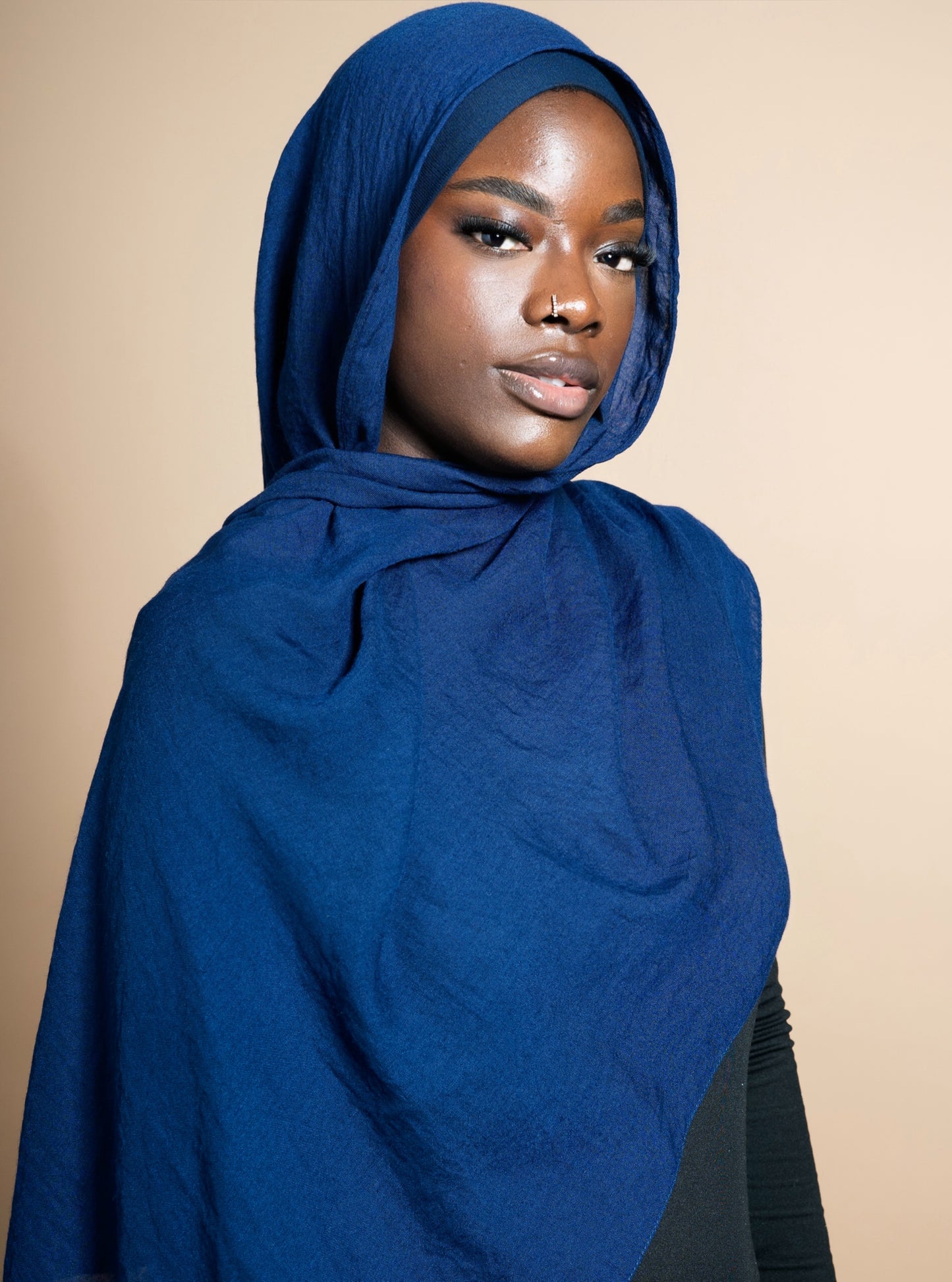 Femme portant un hijab bleu marine en tissu léger et texturé, parfait pour un look modeste et naturel. Matière Coton et Modal. Idéal pour un hijab quotidien confortable et élégant. Fond beige uni.