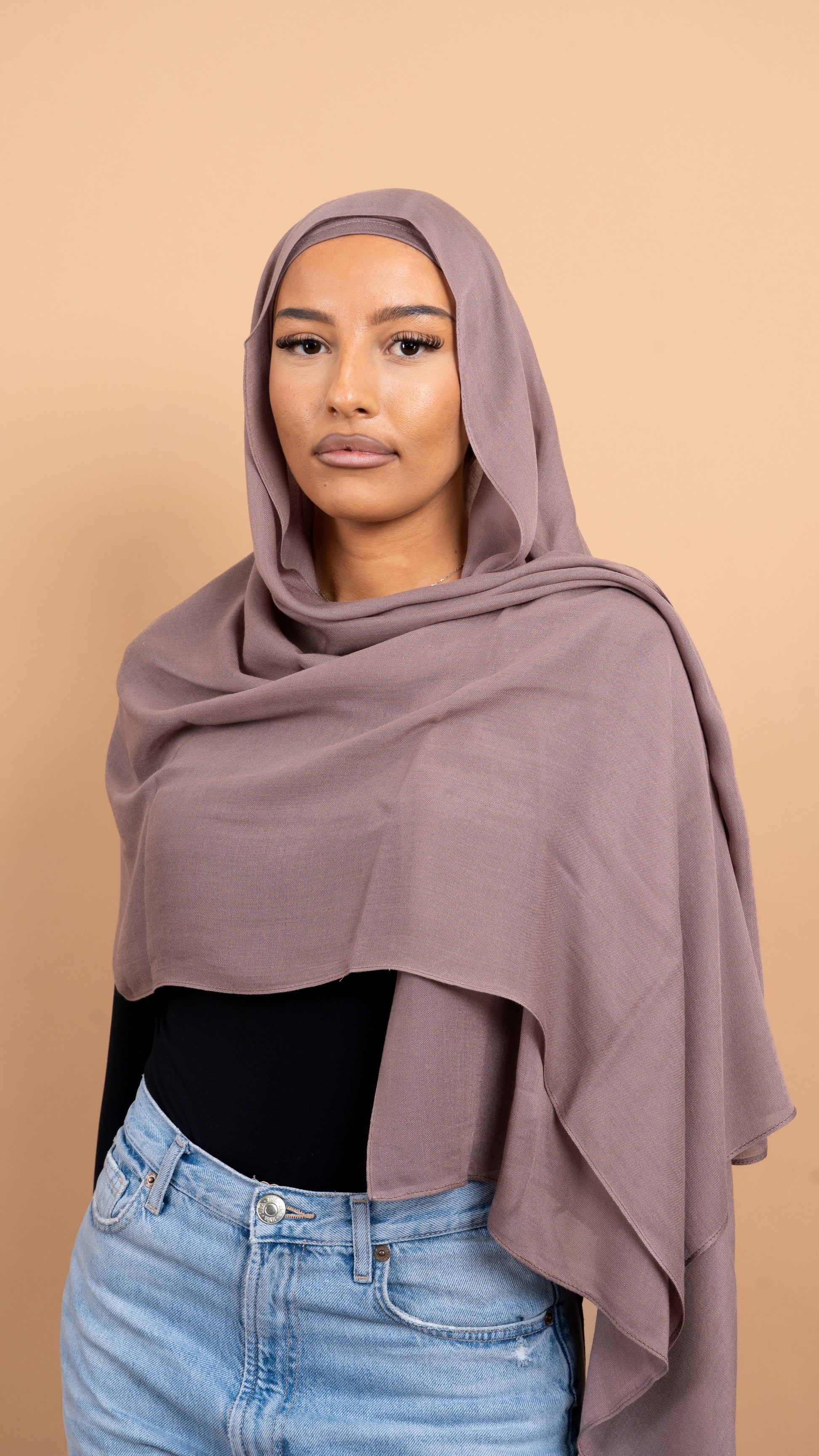 Femme maghrébine voilée avec un foulard en modal de couleur taupe. Photo sur fond beige uni.
