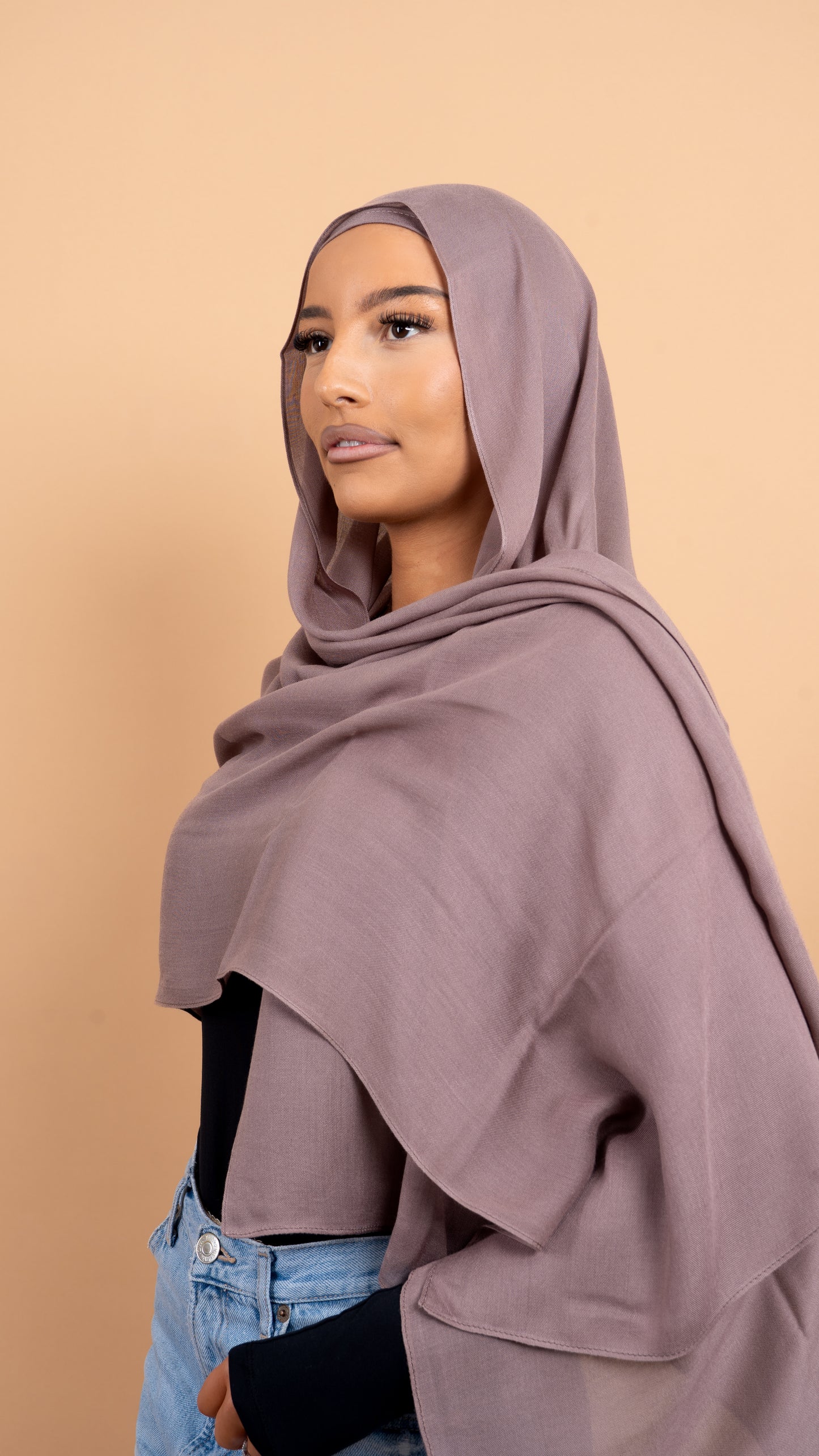 Femme maghrébine voilée avec un foulard en modal de couleur taupe. Photo sur fond beige uni.