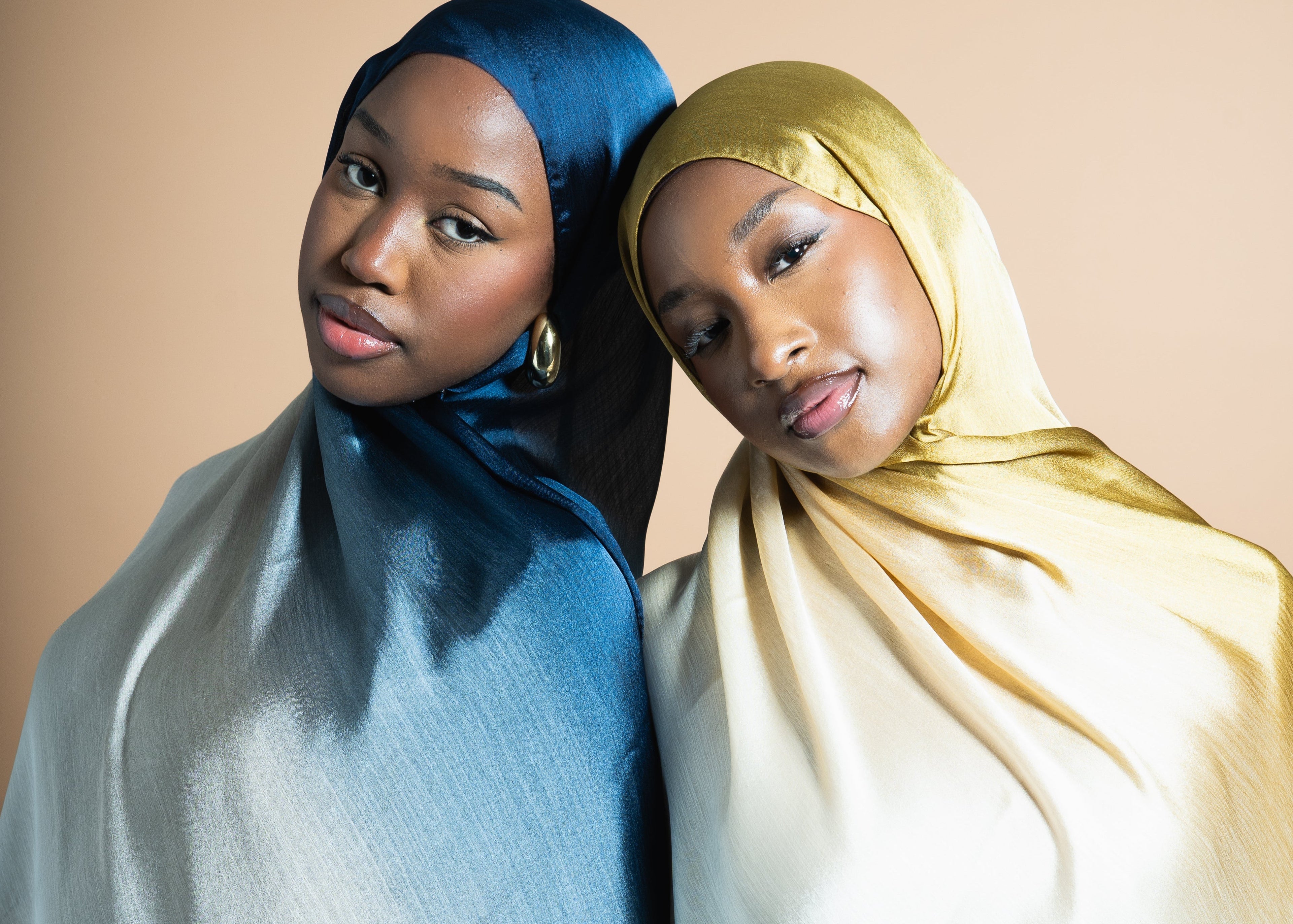 Duo de 2 femmes avec un foulard dégradé chacune. À gauche, la femme porte un foulard satiné bleu à effet dégradé, du marine au gris clair. À droite, la femme porte un foulard satiné jaune doré à effet dégradé, du jaune au beige clair. Fond beige uni.