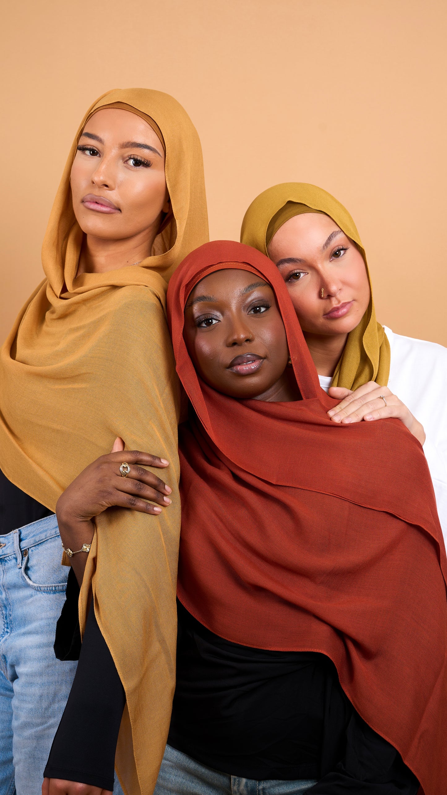Trois femmes voilées de carnation différentes. Une femme noire, blanche et maghrébine. Avec chacune un voile orange, vert olive et jaune moutarde. Photo sur fond beige uni.
