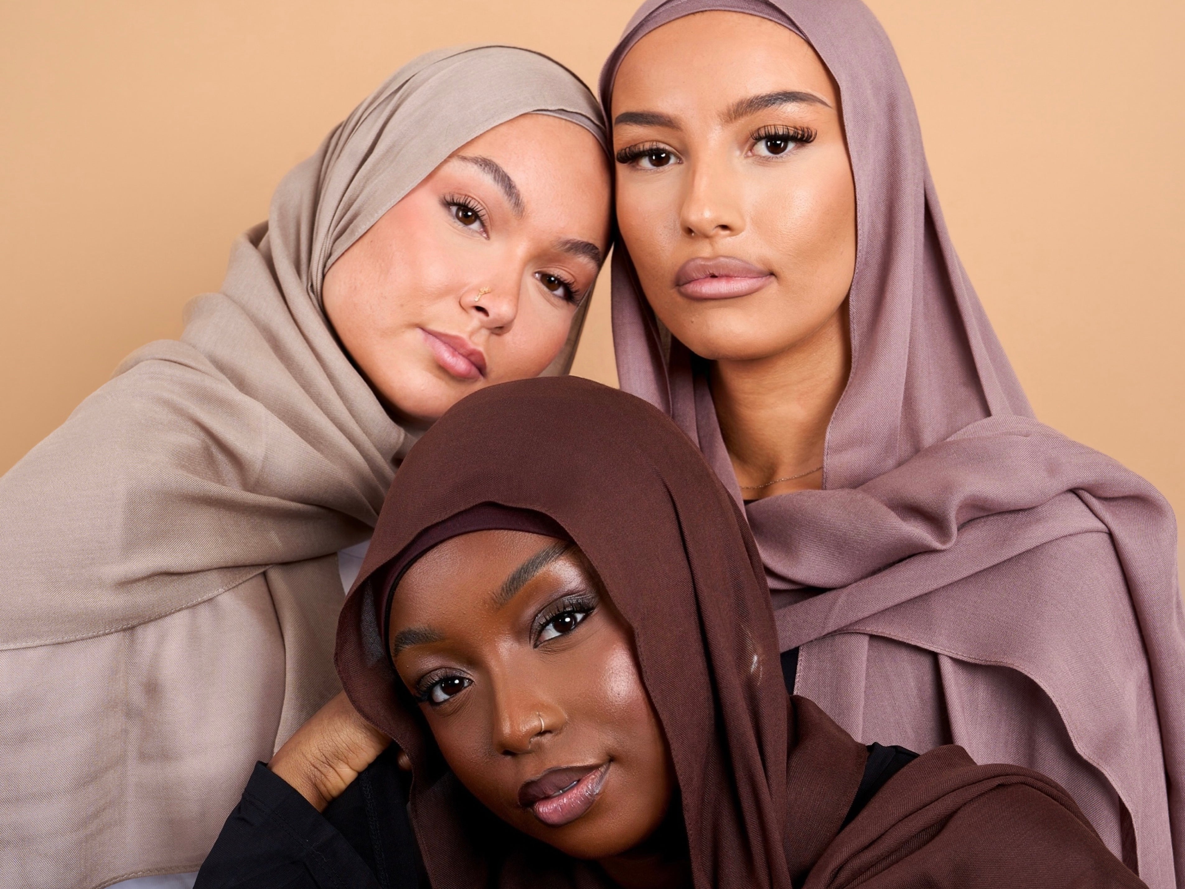 Trois femmes voilées de carnation différentes. Une femme noire, blanche et maghrébine. Avec chacune un voile marron, taupe et gris. Photo sur fond beige uni.